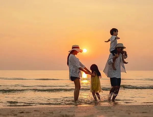 A family paddling at the water’s edge at sunset, one child on their father’s shoulders